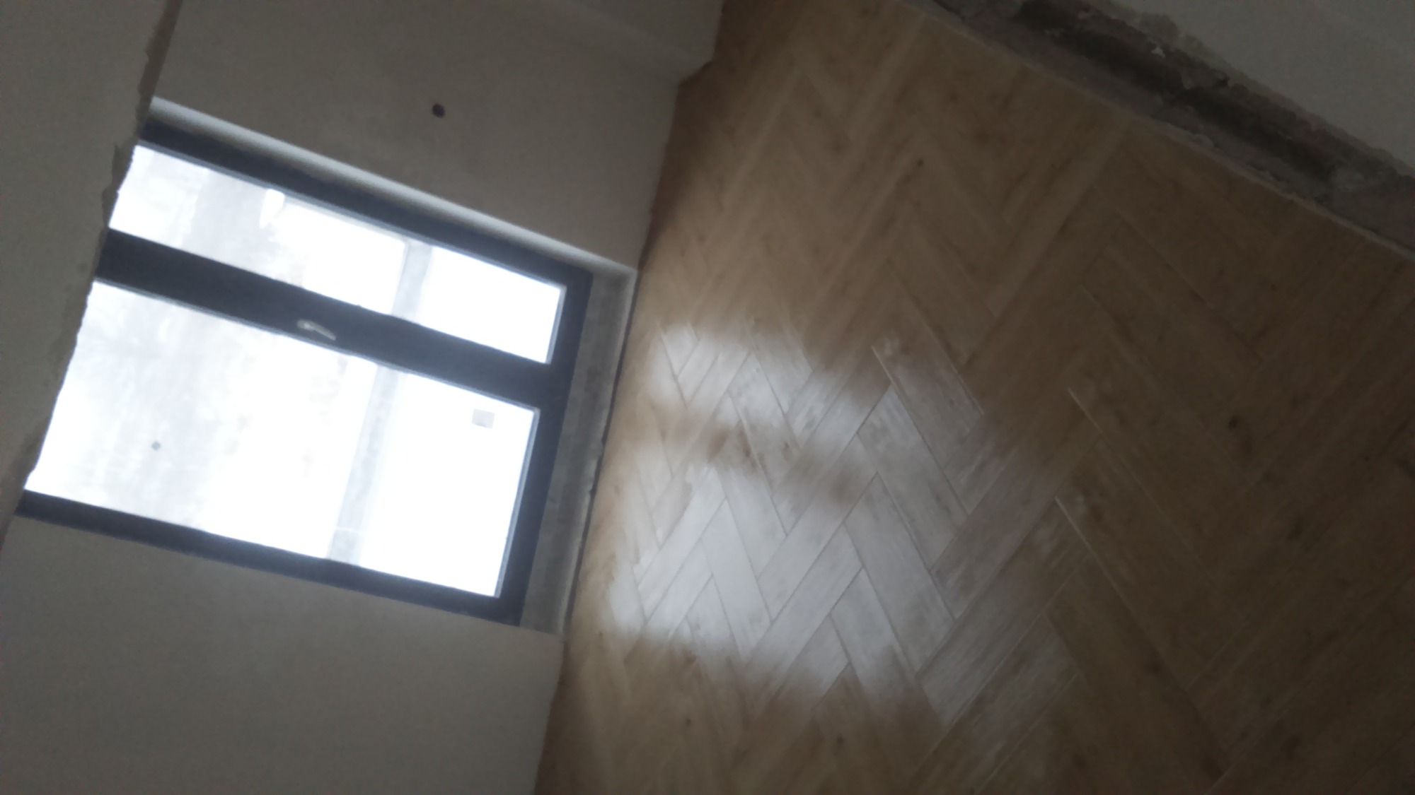 Jasne panele ułożone w jodełkę w nowym pomieszczeniu, widok z góry na podłogę i okno z czarną ramą.