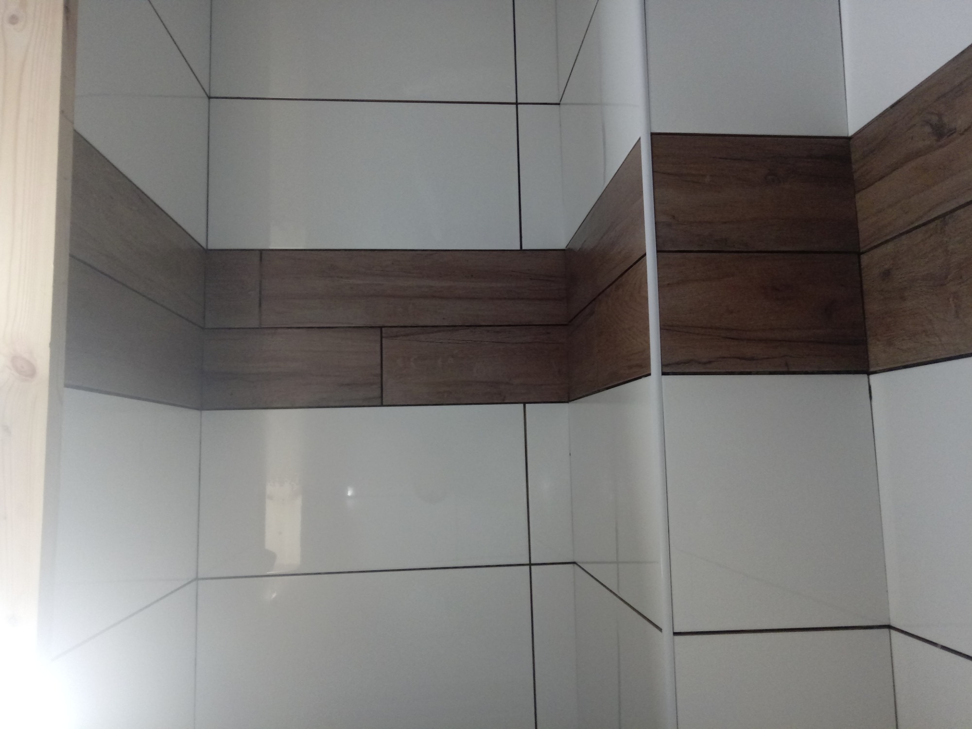 Fragment wykończonej łazienki z białymi płytkami i pasem dekoracyjnym z płytek imitujących drewno, widoczne narożniki i fugi.