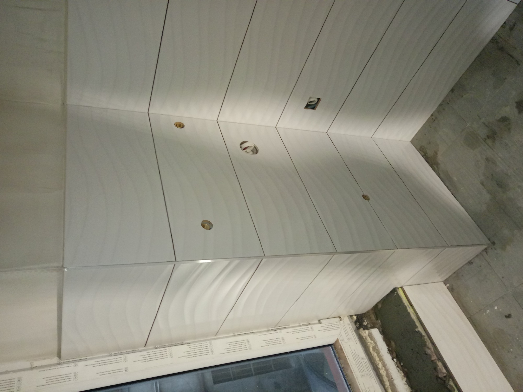 Ściana wyłożona białymi, podłużnymi płytkami z delikatnym wzorem fal, widoczne otwory na oświetlenie, niedokończone wykończenie przy oknie.