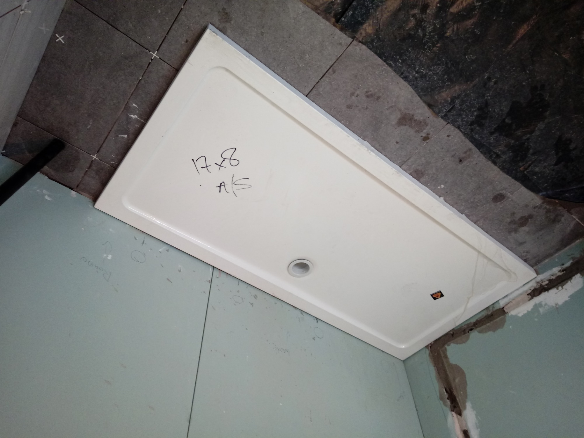 Biały brodzik z odręcznym napisem '17x8 A/S' leży na szarych płytkach podłogowych w trakcie prac wykończeniowych w łazience, ściany pokryte płytami kartonowo-gipsowymi.