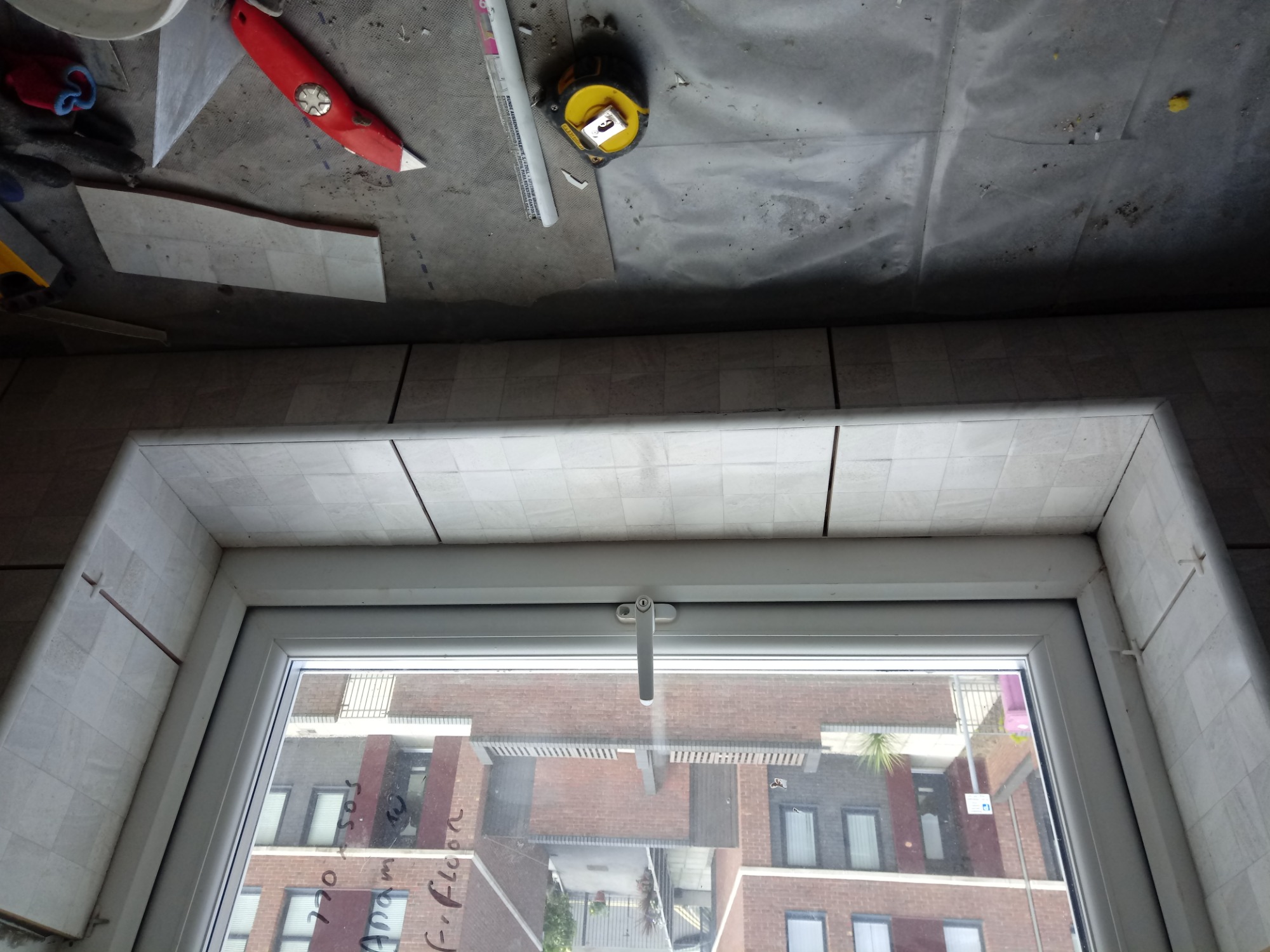 Okładzina okienna z płytek ceramicznych w trakcie prac, widoczne narzędzia: nóż, miarka, ołówek, w tle budynek mieszkalny odbity w szybie okna.