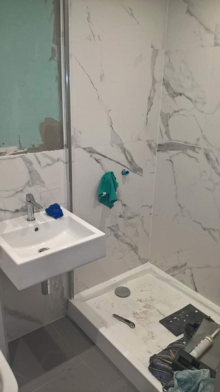 Niedokończona łazienka z białymi płytkami imitującymi marmur, widoczna kabina prysznicowa z narzędziami, umywalka z baterią, oraz fragment ściany z widocznymi śladami po pracach wykończeniowych.