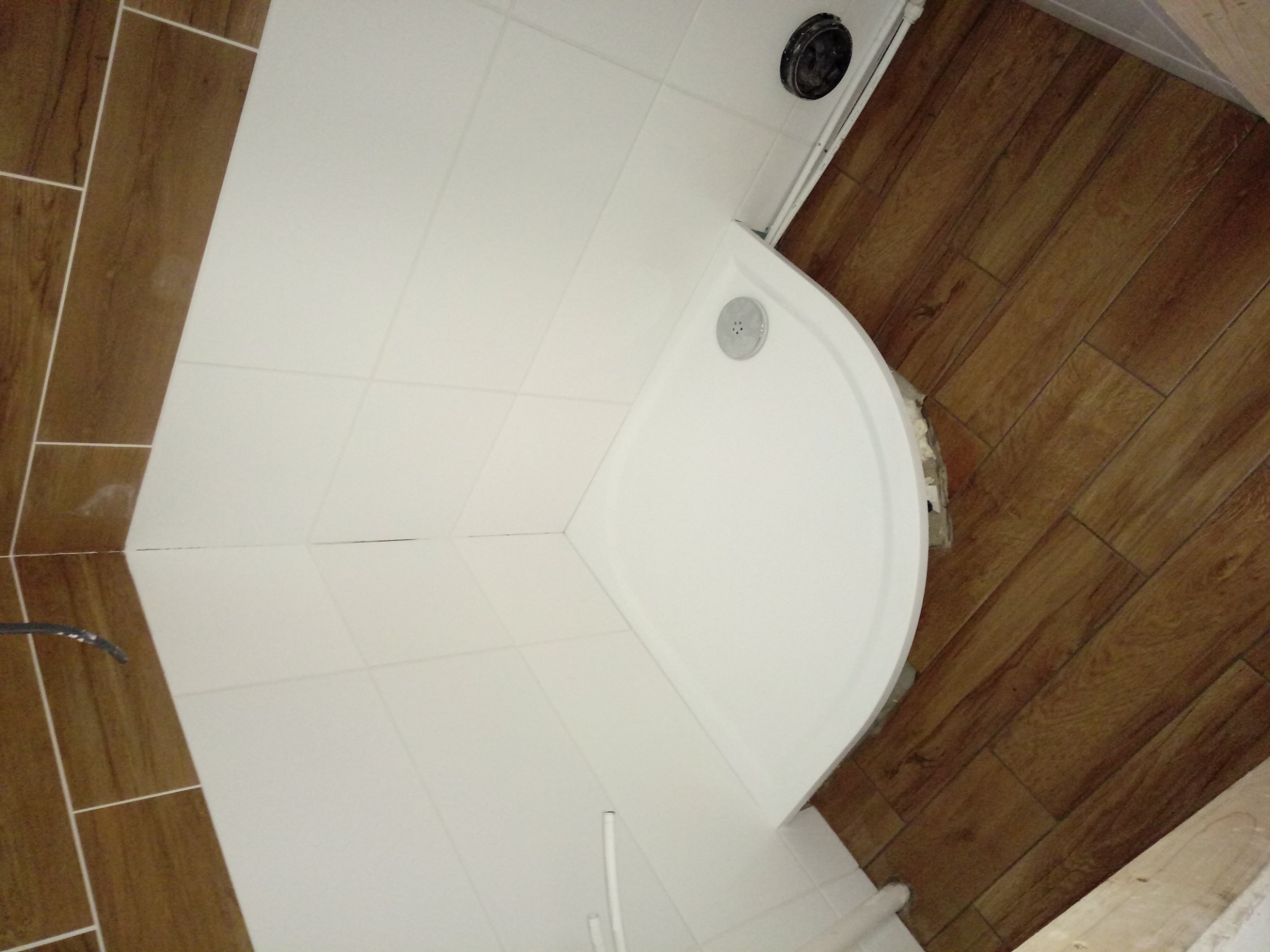 Narożna kabina prysznicowa z białym brodzikiem, białe płytki na ścianie i brązowe płytki imitujące drewno na podłodze, widok z góry.