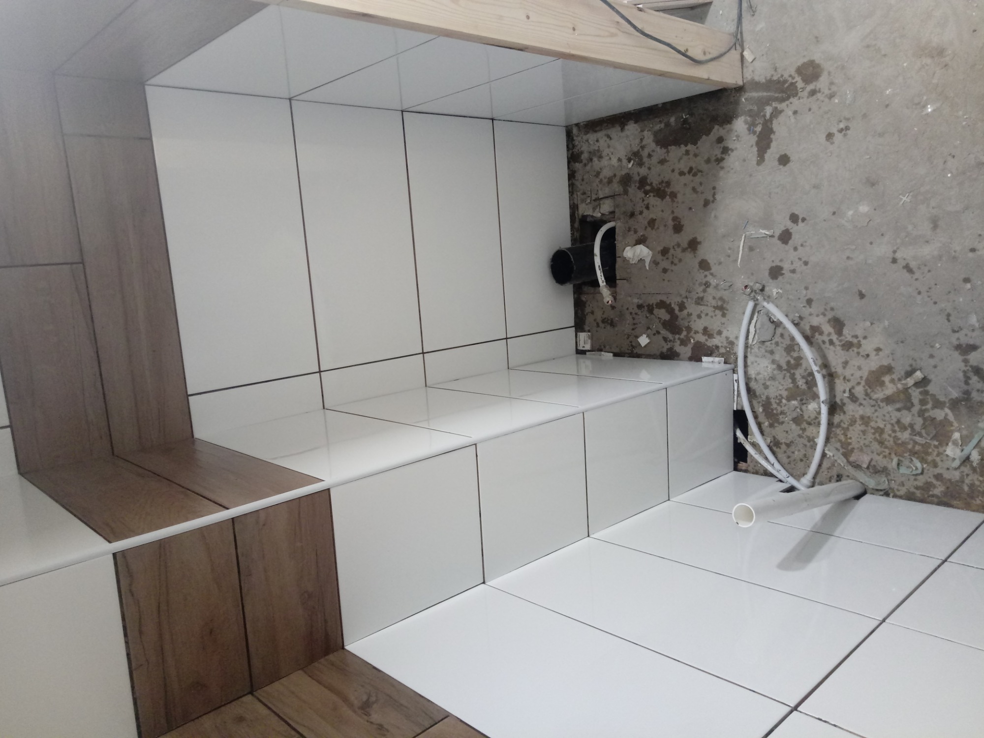 Wnętrze łazienki w trakcie remontu: białe i drewnopodobne płytki ułożone na ścianach i podłodze, widoczne rury i surowa ściana w tle.