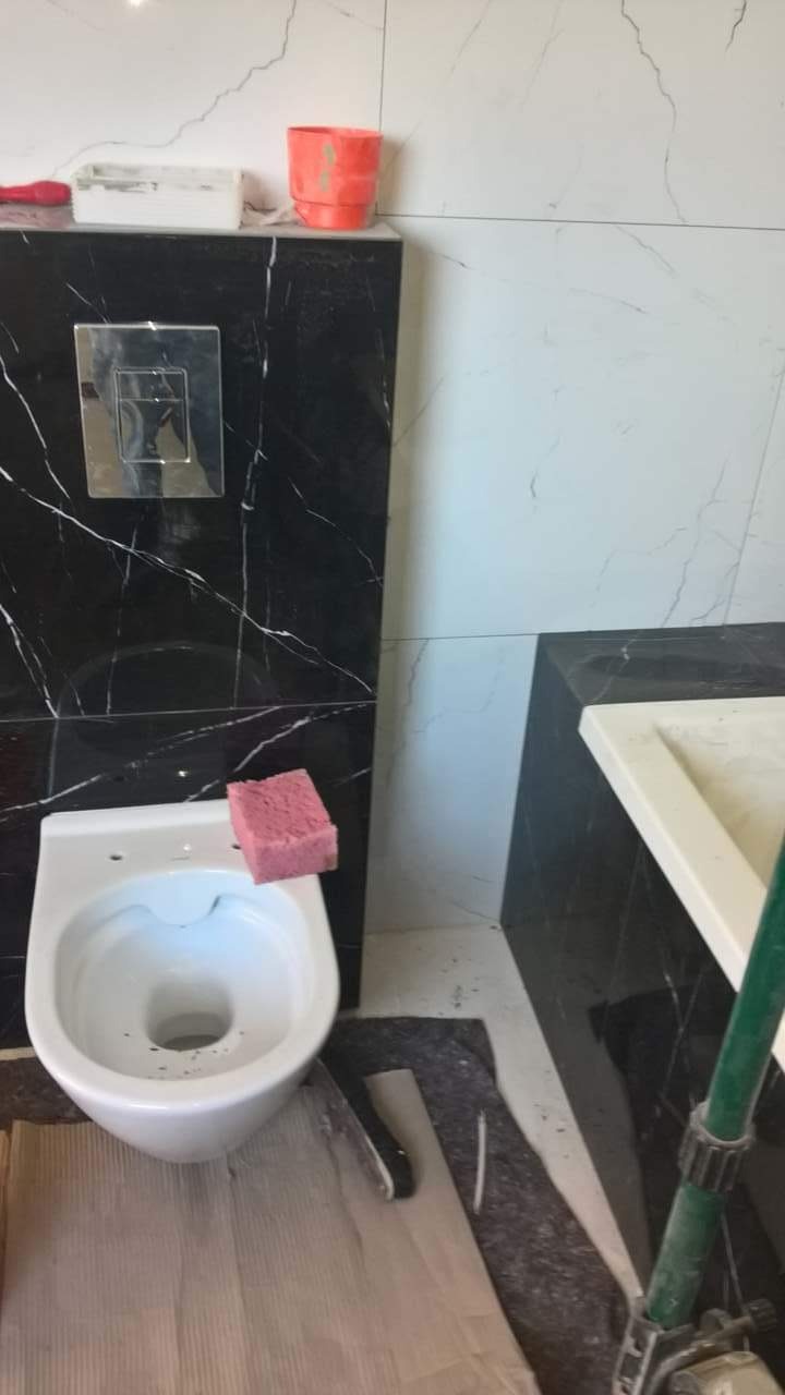 Biała miska WC wisząca z różową gąbką na krawędzi, fragment czarnej marmurowej ściany z przyciskiem spłukującym, białe marmurowe płytki na ścianie, elementy instalacji hydraulicznej na podłodze...