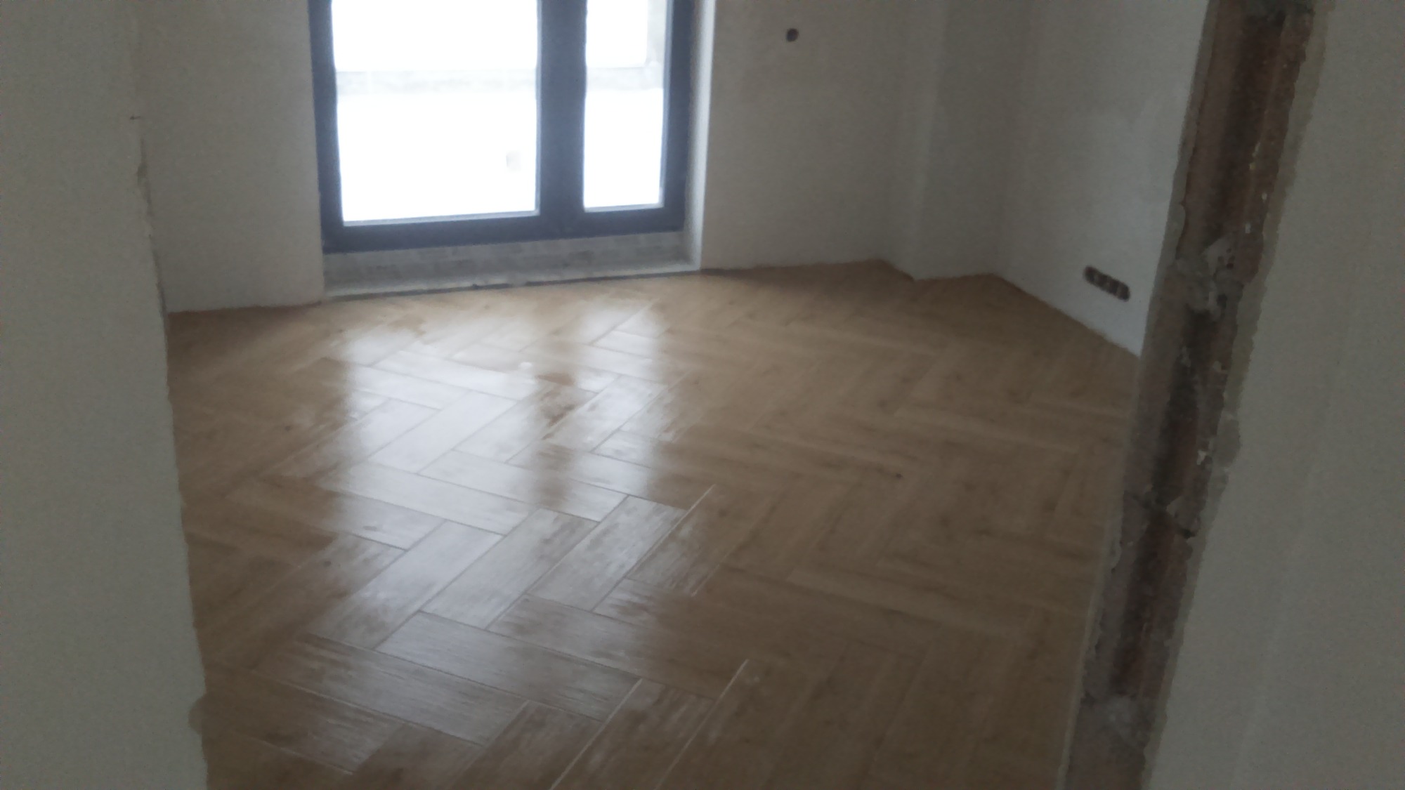 Ułożona podłoga z paneli w jodełkę w pustym pokoju z oknem i surowymi ścianami, widoczne niedokończone wykończenia i wystające kable elektryczne.