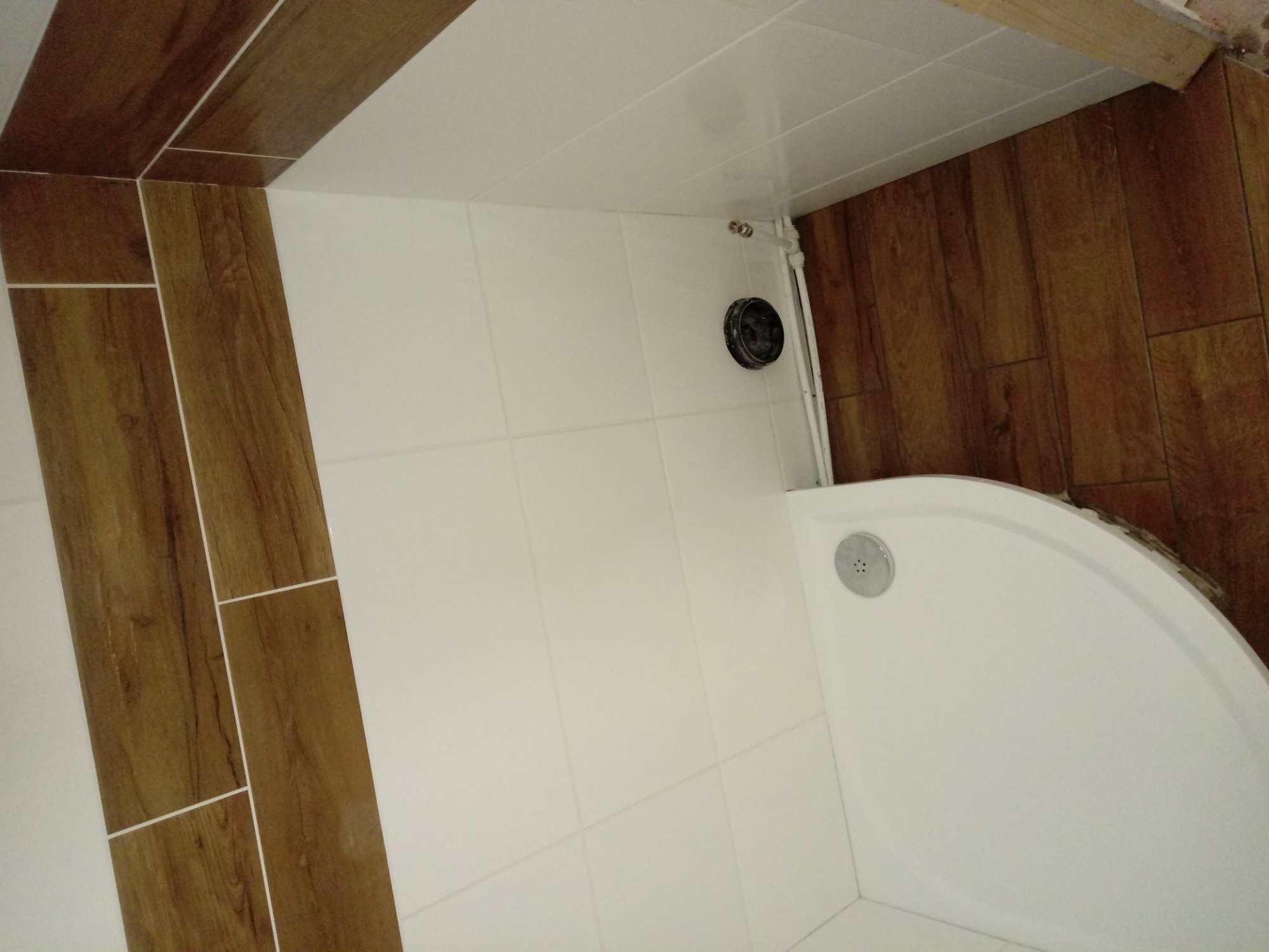 Narożna kabina prysznicowa z odpływem, białe płytki na ścianach, brązowe płytki imitujące drewno na ścianach i suficie, widoczne rury doprowadzające wodę.