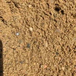 Bliskie ujęcie gruboziarnistego materiału sypkiego o barwie piaskowej, z widocznymi drobnymi kamykami i ciemniejszymi ziarnami, oświetlone z góry, z cieniem po lewej stronie kadru.