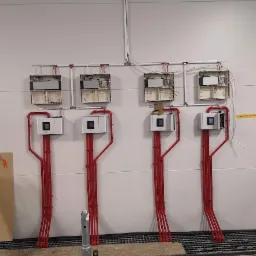 Instalacja czterech identycznych systemów z białymi obudowami i czerwonymi rurami na białej ścianie, widoczne otwarte skrzynki elektryczne powyżej, drabina i fragmenty podłogi w tle.