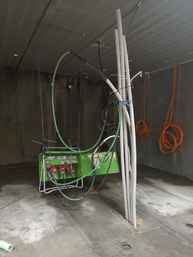 Zielona skrzynka elektryczna z czerwonymi gniazdami zawieszona na linach w surowym wnętrzu, obok wiązka białych rur i zwisające pomarańczowe kable.