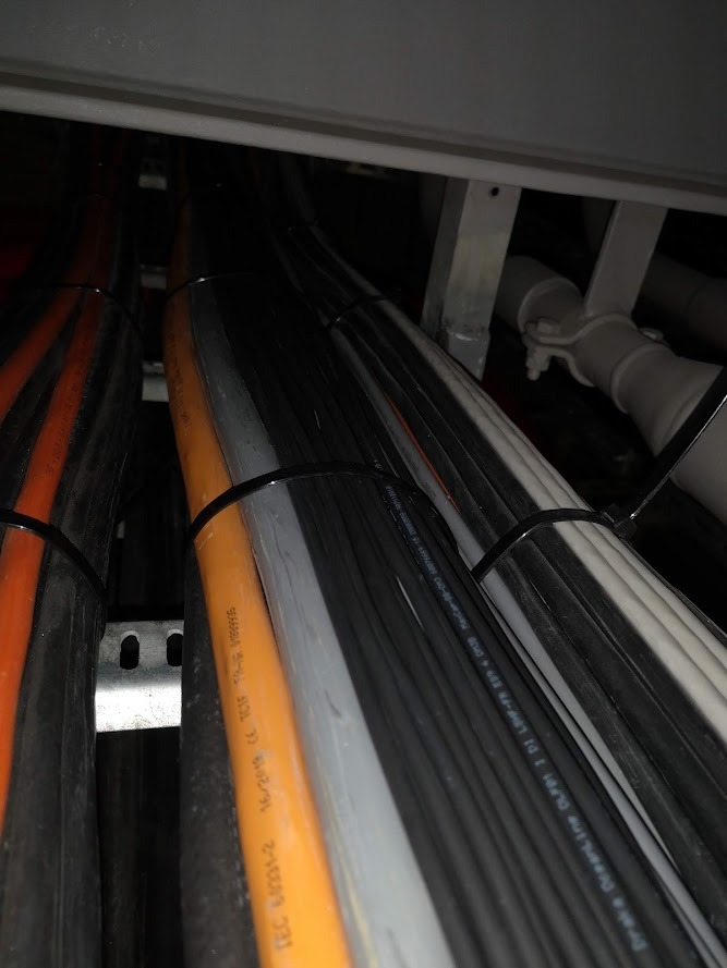 Wiązka przewodów elektrycznych w różnych kolorach (pomarańczowy, szary, czarny, biały) spięta czarnymi opaskami zaciskowymi, ułożona na metalowej konstrukcji wewnątrz szafy rozdzielczej lub koryta...