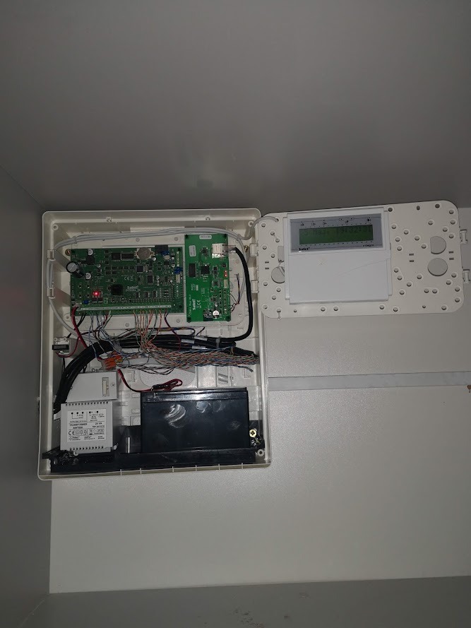Otwarta obudowa centrali alarmowej z widoczną elektroniką, akumulatorem i przewodami, zamontowana na białej ścianie.