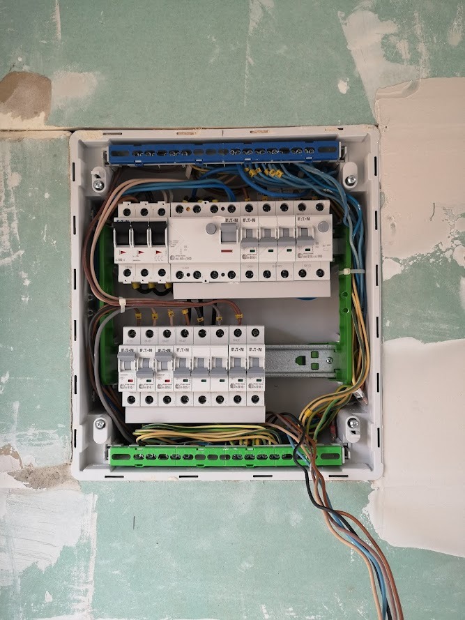 Otwarta skrzynka elektryczna z zainstalowanymi bezpiecznikami i podłączonymi przewodami, zamontowana w ścianie pokrytej gładzią gipsową przed malowaniem.