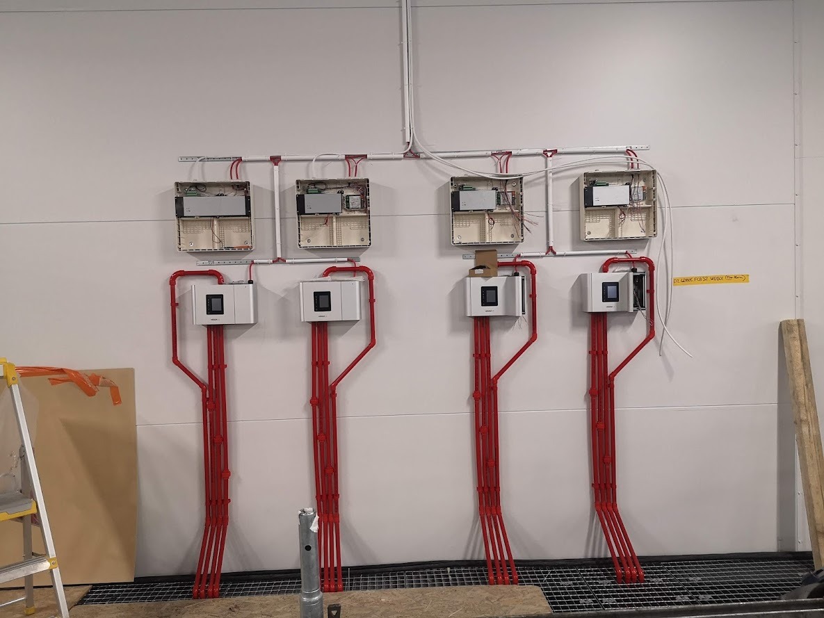 Instalacja czterech identycznych systemów z białymi obudowami i czerwonymi rurami na białej ścianie, widoczne otwarte skrzynki elektryczne powyżej, drabina i fragmenty podłogi w tle.