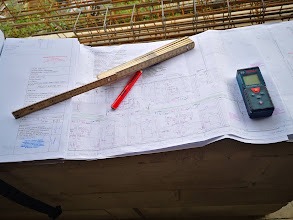 Plan techniczny z naniesionymi pomiarami, leżący na surowym drewnie, obok składana miarka, czerwony długopis i laserowy miernik odległości Bosch.