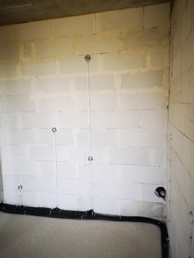 Surowy stan instalacji elektrycznej w pomieszczeniu: przewody w peszlu ułożone wzdłuż ściany z bloczków, widoczne puszki pod gniazdka i wypusty kabli oświetleniowych.