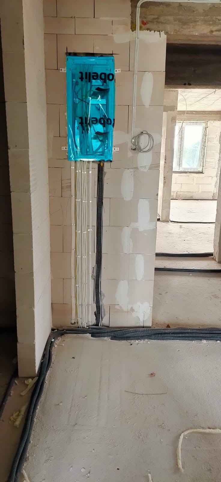 Rozdzielnica elektryczna tymczasowo zabezpieczona folią, z wyprowadzonymi przewodami w peszlach, zamontowana na surowej ścianie z bloczków betonowych w trakcie prac instalacyjnych.