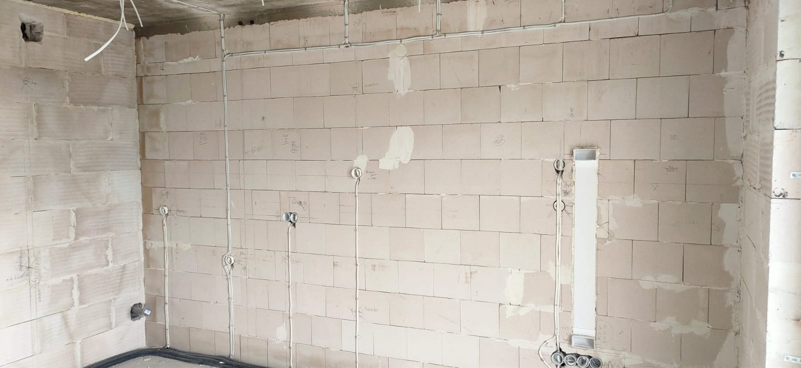 Surowe ściany z betonu komórkowego w trakcie instalacji elektrycznej, widoczne kable w peszlach, puszki pod gniazdka i włączniki, oraz oznaczenia na ścianach.