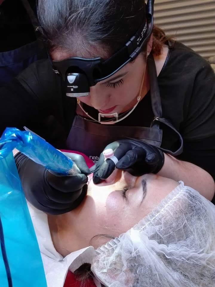 Kobieta w czarnych rękawiczkach wykonuje zabieg pigmentacji ust klientki, która ma założony czepek ochronny na włosach. Używane jest specjalistyczne oświetlenie czołowe.