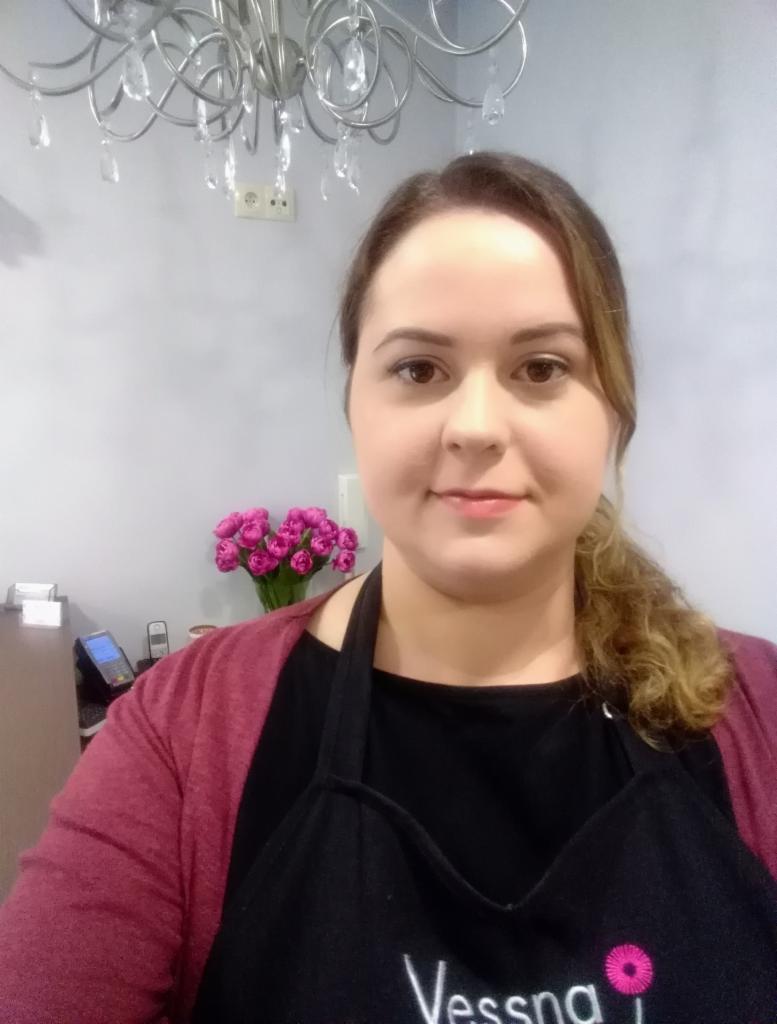 Portret kobiety w czarnym fartuchu z logo, w tle bukiet różowych tulipanów i kryształowy żyrandol w salonie.