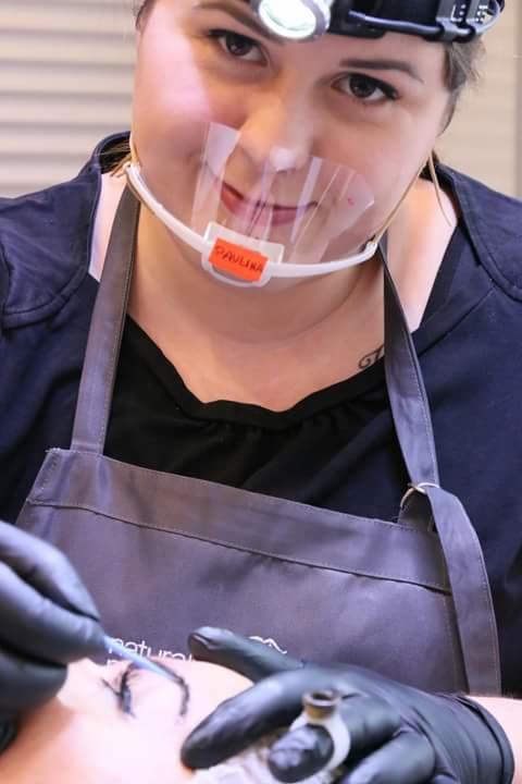 Kosmetolog w czarnych rękawiczkach i przezroczystej osłonie z napisem 'PAVLINA' wykonuje makijaż permanentny brwi klientce.