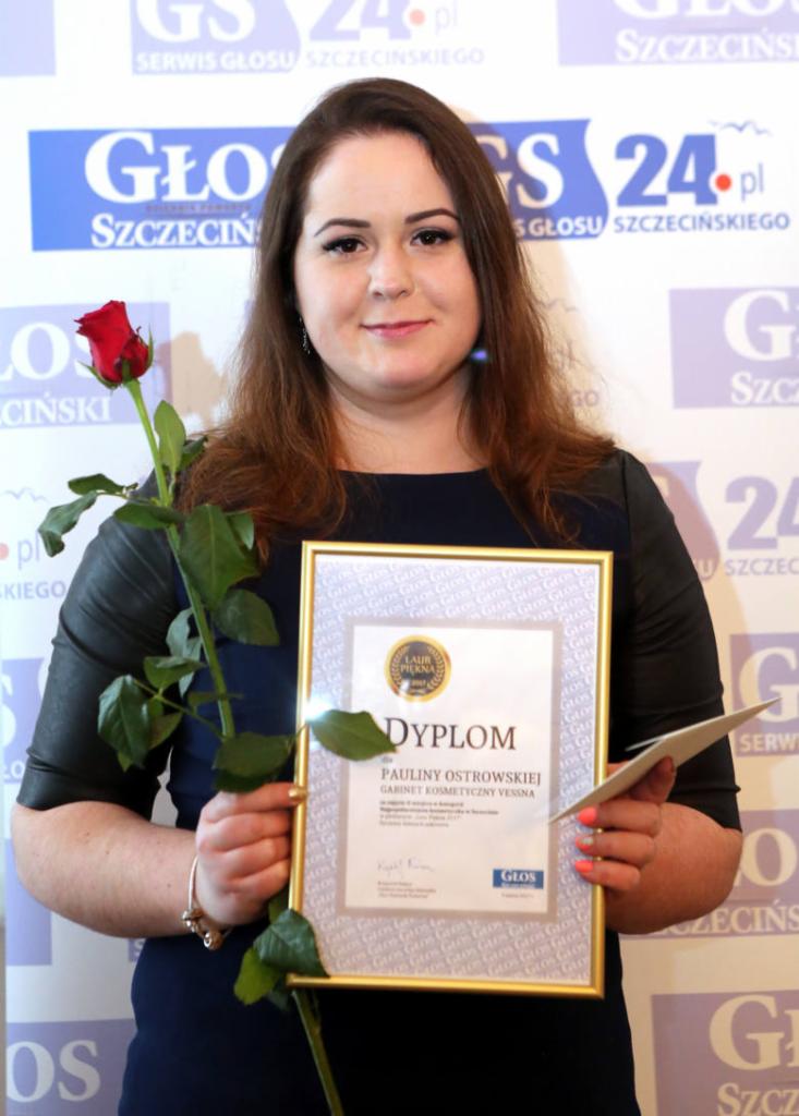Uśmiechnięta kobieta trzymająca dyplom 'Laur Piękna' dla Pauliny Ostrowskiej, Gabinet Kosmetyczny Vessna, z czerwoną różą w drugiej ręce, na tle logo Głos Szczeciński.