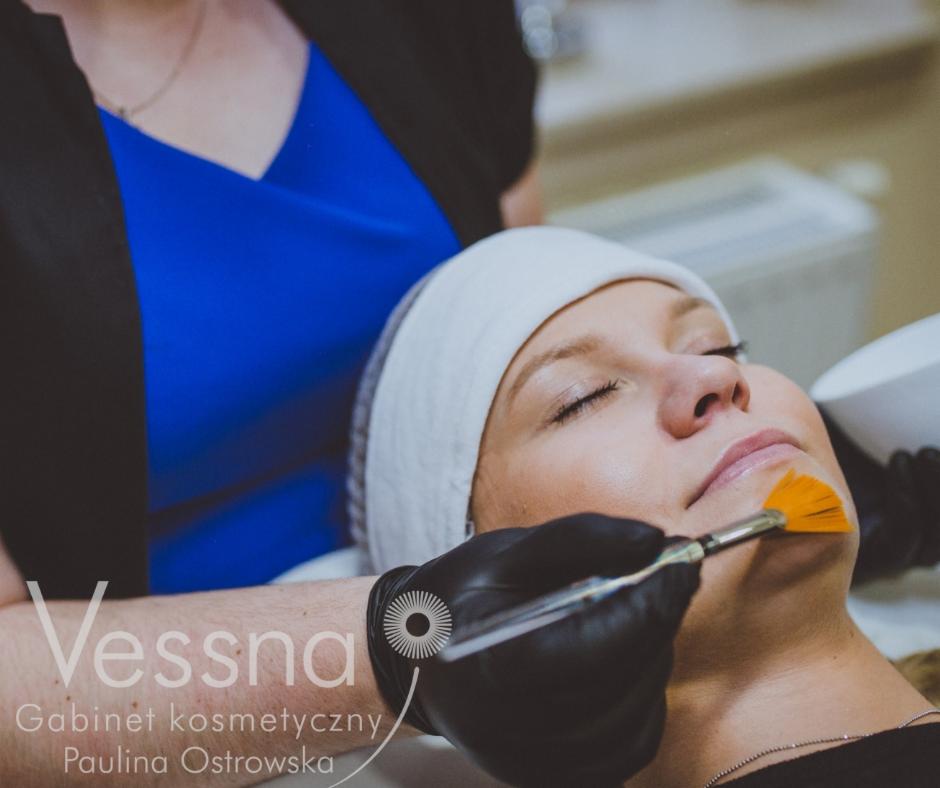 Klientka z zamkniętymi oczami podczas nakładania pomarańczowej maski pędzlem przez kosmetyczkę w czarnych rękawiczkach, biała opaska na włosach.