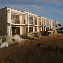 Nowoczesne domy w zabudowie szeregowej z drewnianą elewacją w trakcie budowy, widoczne materiały budowlane i samochód dostawczy na placu budowy.