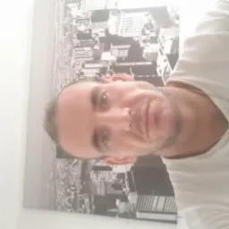 Mężczyzna w białej koszulce na tle czarno-białej grafiki z panoramą miasta. Ujęcie z bliska, widoczna część ramienia i szyi. Oświetlenie naturalne, jasne tło.