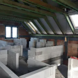 Budowa ścian działowych z białych bloczków na poddaszu z oknami dachowymi, widoczne drewniane elementy konstrukcyjne dachu i fragmenty ścian z cegły.