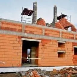 Budowa domu z czerwonej cegły ceramicznej w trakcie wznoszenia ścian, widoczne otwory okienne i drzwiowe, oraz fragmenty rusztowań na dachu.