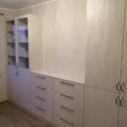 Zabudowa meblowa pod wymiar w jasnej kolorystyce z szufladami i szafkami, widoczna na tle ściany i podłogi imitującej drewno.