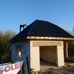 Pracownik montuje czarną dachówkę na nowym dachu budynku w trakcie budowy, widoczny fragment ściany z białego bloczka i częściowo zamontowana rynna.