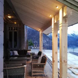 Drewniany taras z meblami rattanowymi i widokiem na góry i jezioro, oświetlony punktowymi lampami sufitowymi.