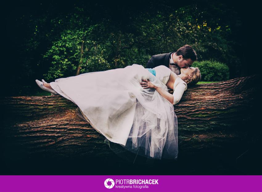 Panna młoda w białej sukni i pan młody w czarnym garniturze całują się, leżąc na powalonym drzewie w lesie, delikatny makijaż panny młodej, ciemnozielone tło.