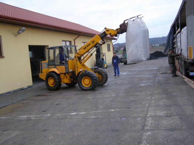 Żółta ładowarka teleskopowa JCB TM200 podnosi duży worek typu big bag z pelletem i umieszcza go na przyczepie ciężarówki, w tle budynek gospodarczy i pracownicy.