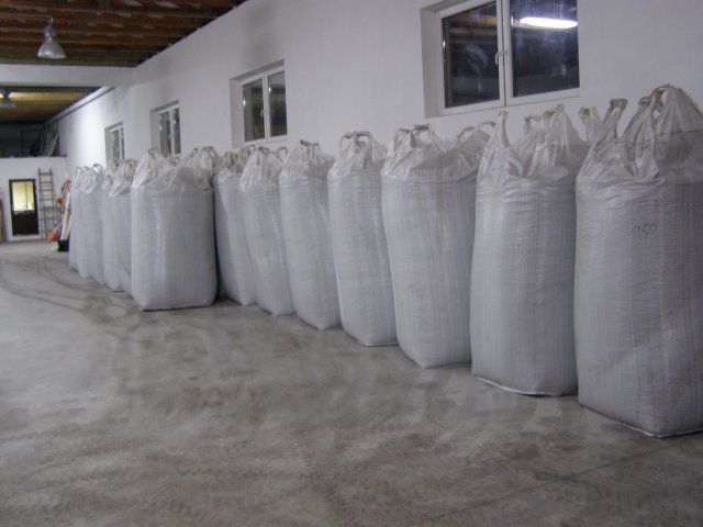 Rząd worków z pelletem ustawionych w magazynie z betonową podłogą i białymi ścianami, widoczne okna i drzwi w tle.