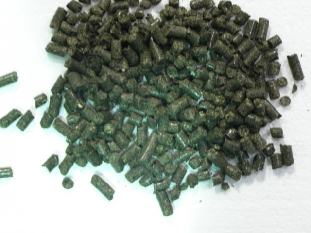 Kupa ciemnych, cylindrycznych peletek na jasnej powierzchni, widoczne drobne włókna w strukturze peletek.