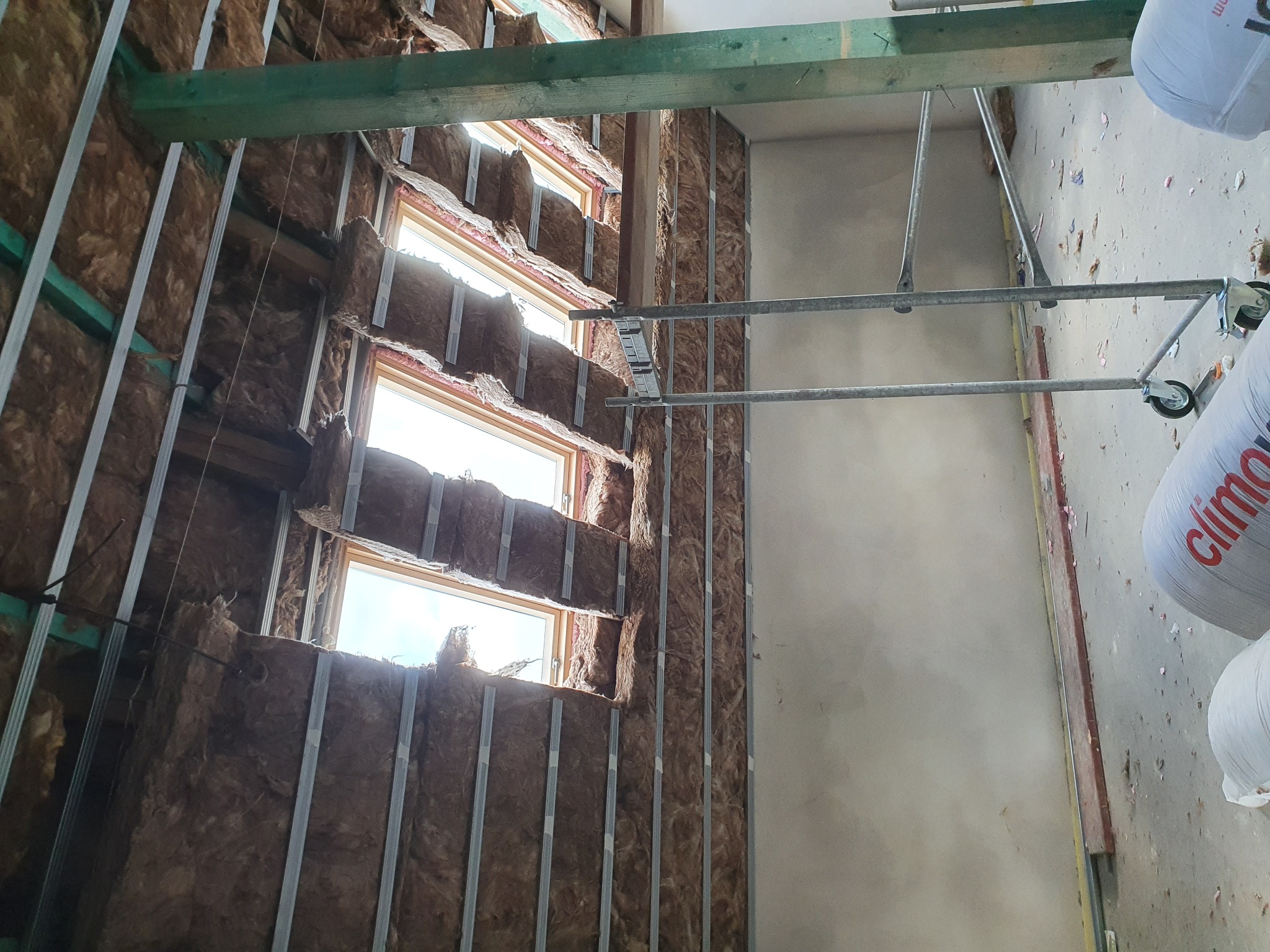 Ocieplenie poddasza wełną mineralną, widoczne okna dachowe i metalowy stelaż pod płyty gipsowo-kartonowe, częściowo otynkowana ściana, na podłodze worki z wełną.