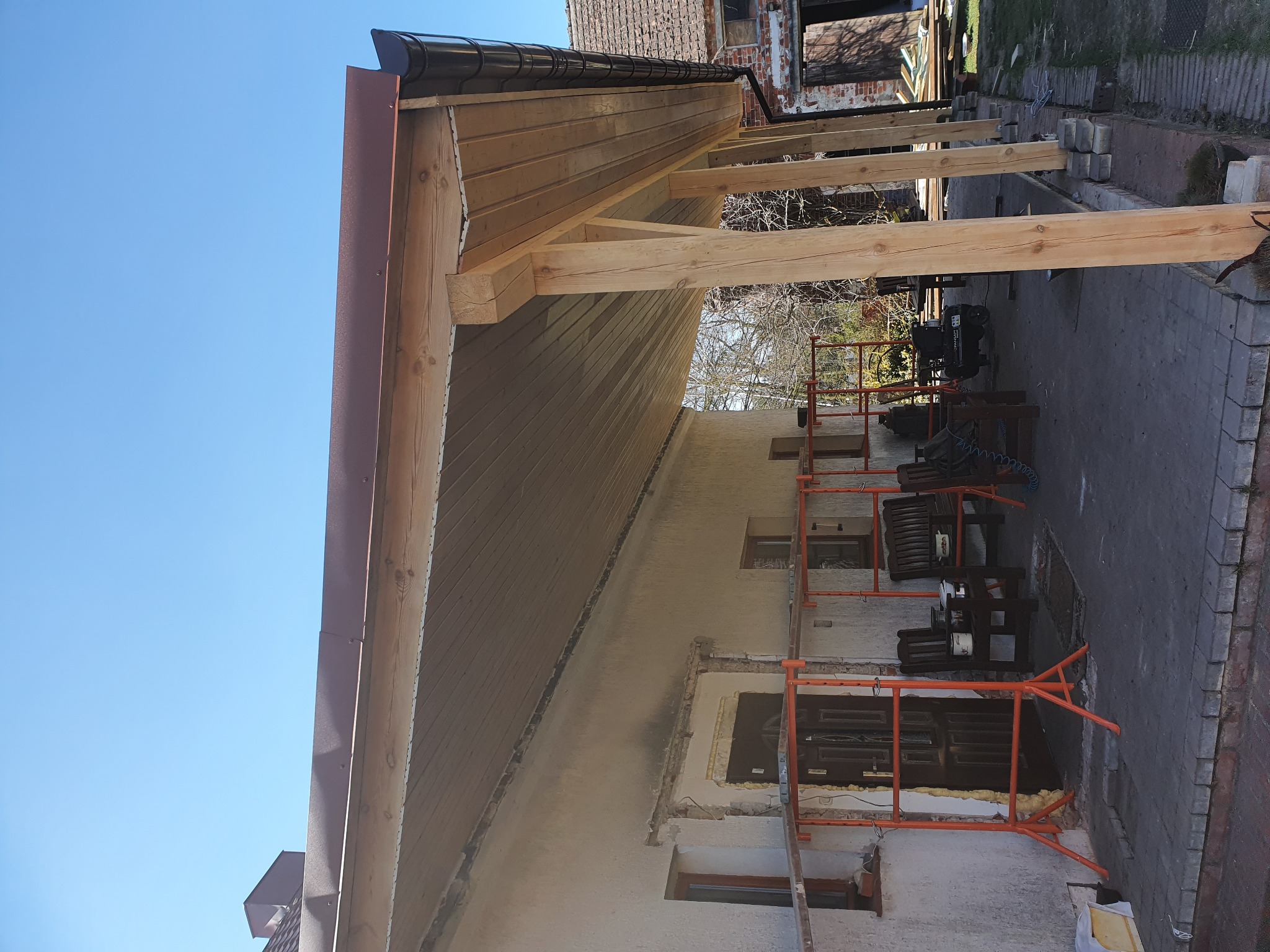 Budowa zadaszenia tarasu z drewnianą konstrukcją i brązowym orynnowaniem, widoczne rusztowanie i przygotowane otwory okienne w ścianie budynku.