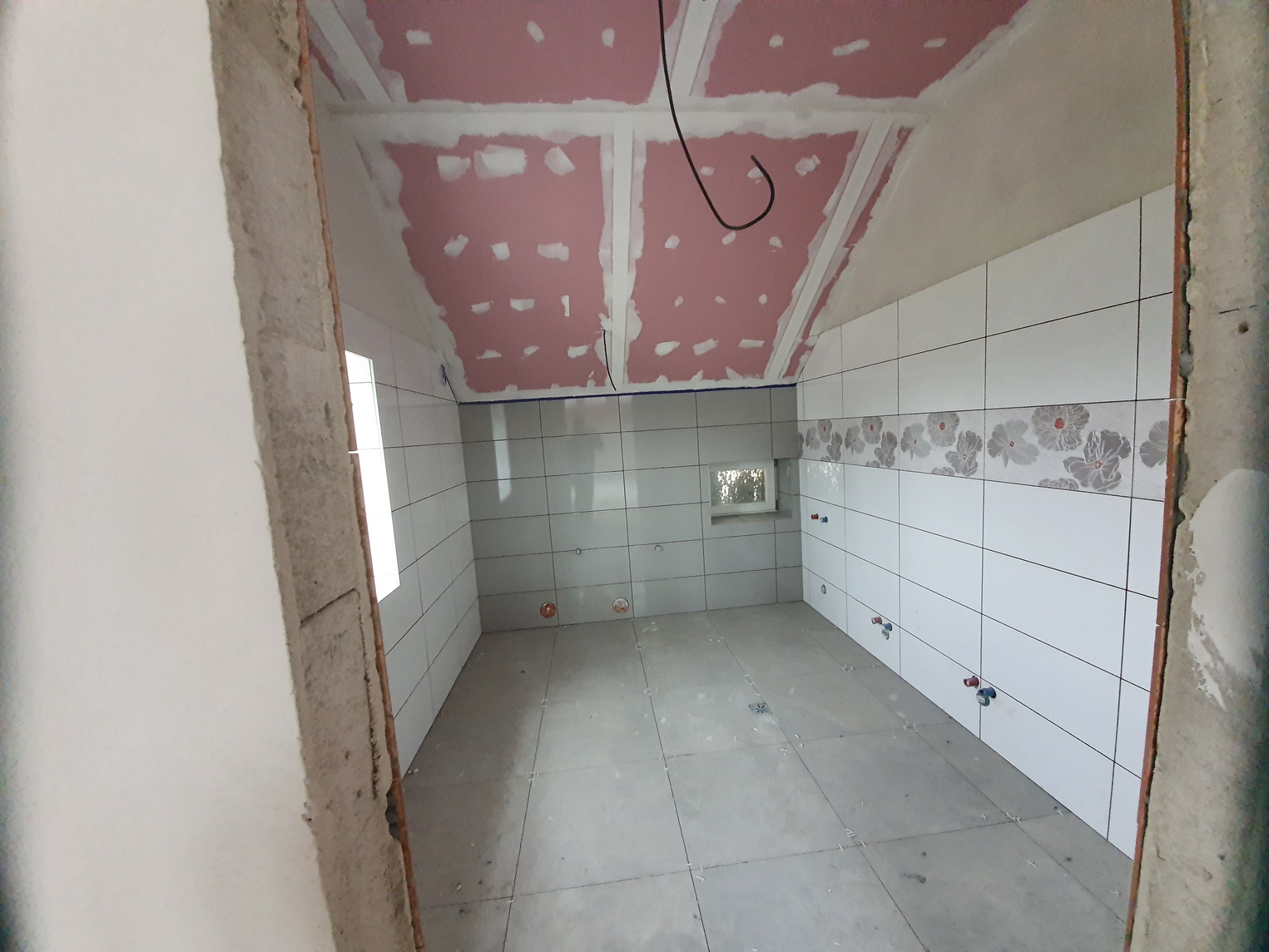 Łazienka w trakcie wykańczania w Szamotułach: sufit z płyt gipsowo-kartonowych po szpachlowaniu, częściowo ułożone białe płytki ścienne z delikatnym kwiatowym wzorem na górze, przygotowane punkty...