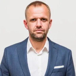 Portret mężczyzny w niebieskiej marynarce i białej koszuli na jasnym tle, z widoczną kieszonką na piersi i zamyślonym wyrazem twarzy.