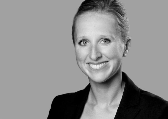 Portret uśmiechniętej kobiety w czarnym żakiecie na szarym tle, czarno-białe zdjęcie.