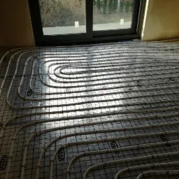 Białe rurki ogrzewania podłogowego ułożone na czarnej siatce, widoczne okno w tle i ściany w odcieniach beżu. Instalacja KISAN.