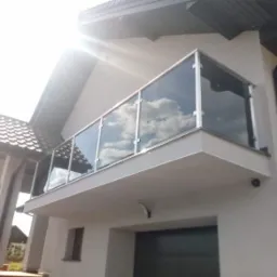 Nowoczesna balustrada szklana na balkonie domu jednorodzinnego, z widocznym odbiciem błękitnego nieba i chmur w szklanych panelach. Minimalistyczny design.