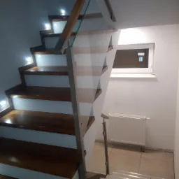 Nowoczesne schody drewniane z podświetleniem LED w stopniach i szklaną balustradą, widok z dołu na górę, z grzejnikiem i oknem w tle