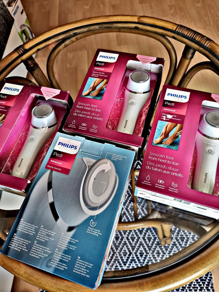 Cztery pudełka z elektrycznymi pilnikami do stóp Philips Pedi Advanced, ułożone na wiklinowym stoliku kawowym z wzorzystym obrusem.