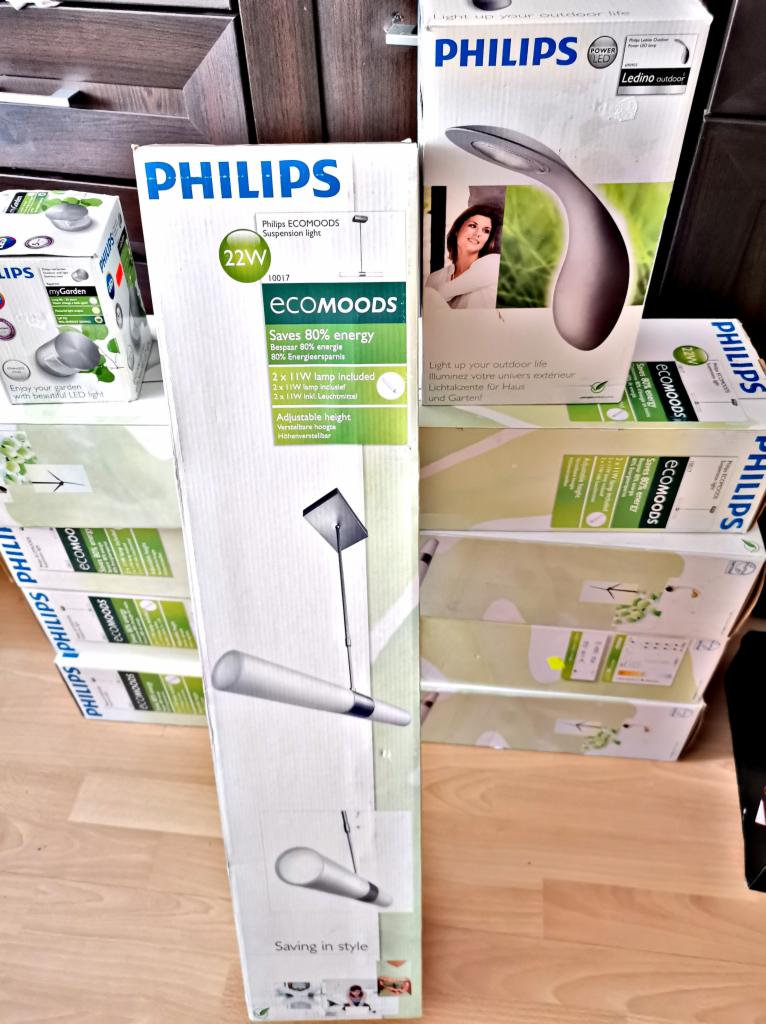 Stos pudełek z lampami Philips ECOMOODS, w tym lampa wisząca i lampa ogrodowa, prezentowane na drewnianej podłodze.