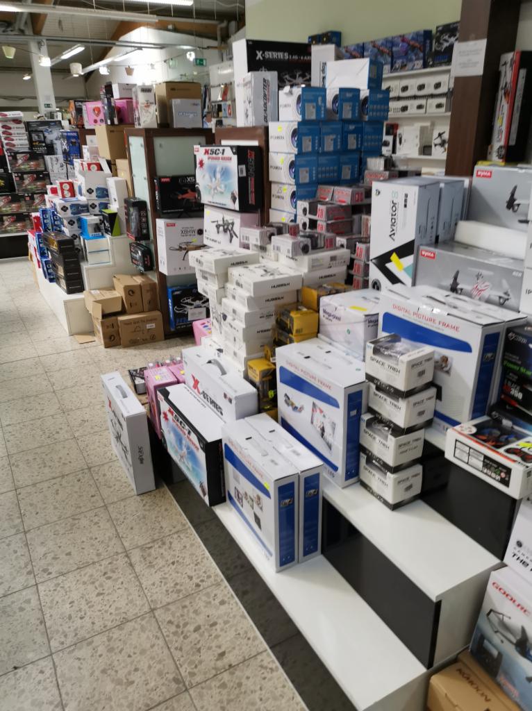 Półka w sklepie wypełniona pudełkami z dronami, kamerami i ramkami cyfrowymi różnych marek, ułożonymi w stosy i rzędy, z widocznym oświetleniem sufitowym i wzorzystą podłogą.