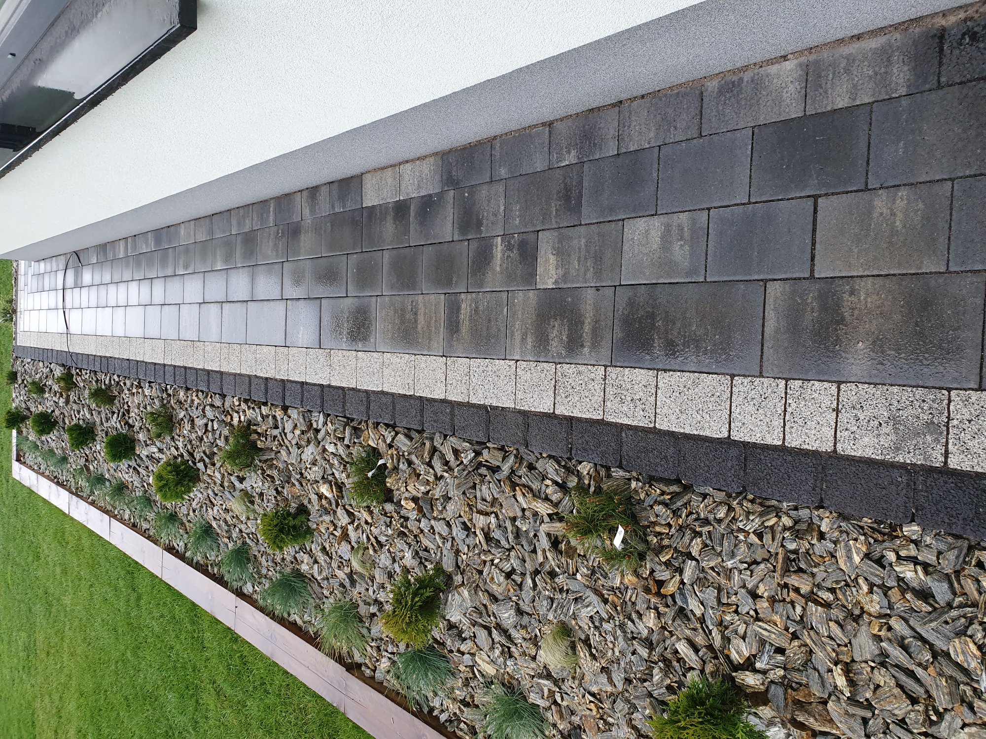 Świeżo ułożona, mokra nawierzchnia z ciemnoszarych kostek brukowych, oddzielona pasem z jasnych kostek od rabaty z korą, niskimi krzewami i trawami ozdobnymi, przylegająca do ściany budynku.